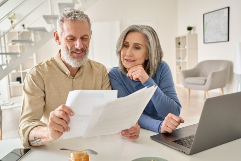A senior couple sit at a laptop comparing multiple documents